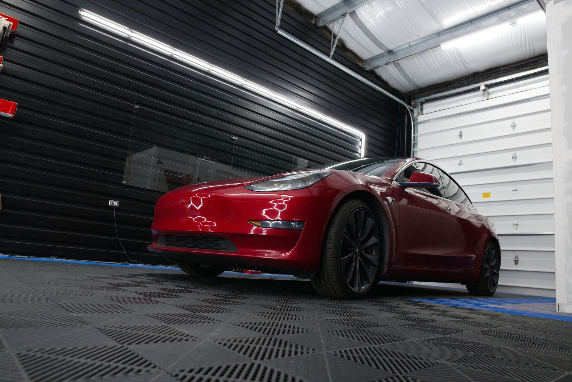 A red Tesla Model 3 parked in a modern garage with black slatted walls and white flooring.