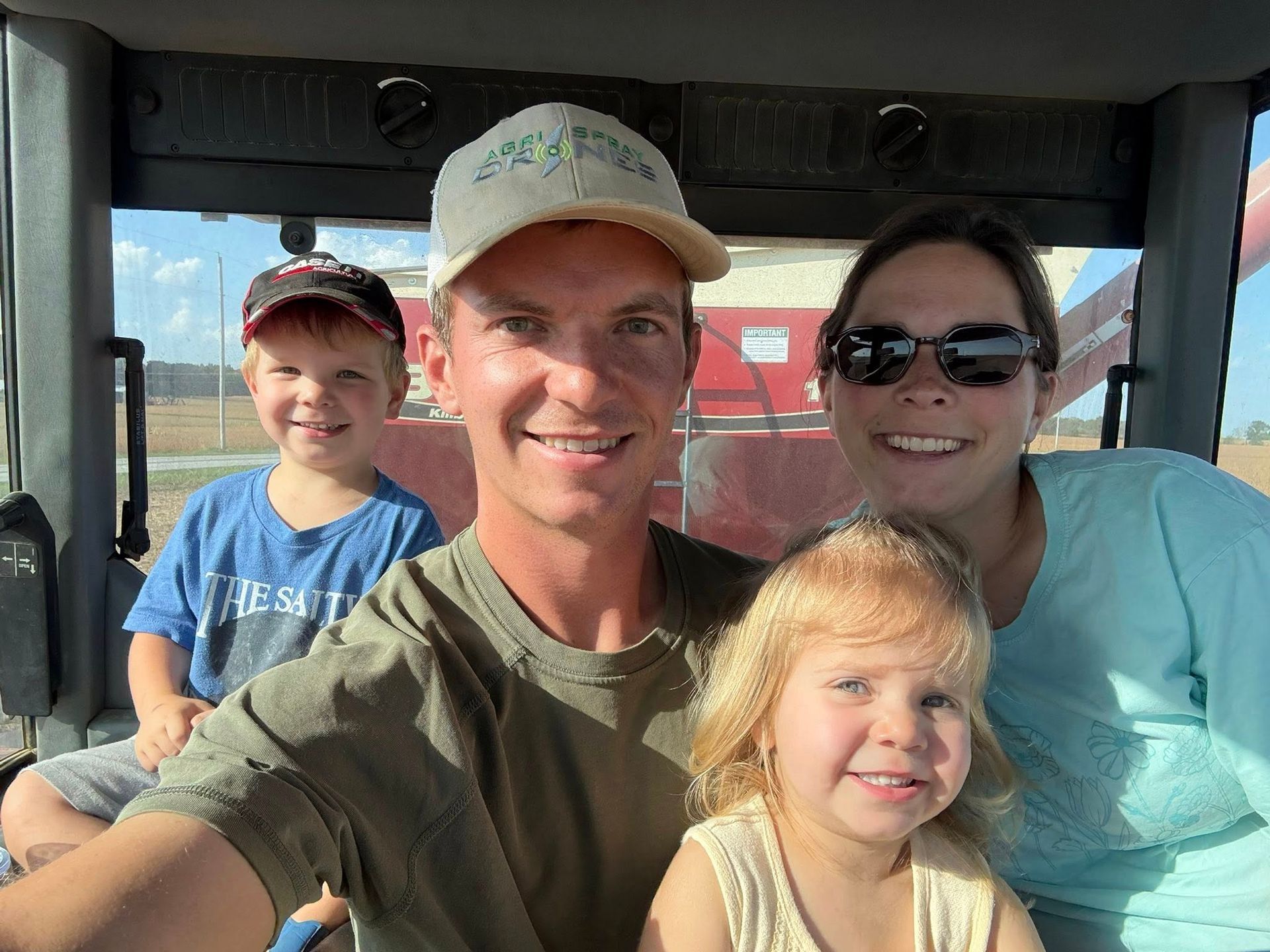 Family in a tractor cab smiles for a selfie; sunny day, field in background.
