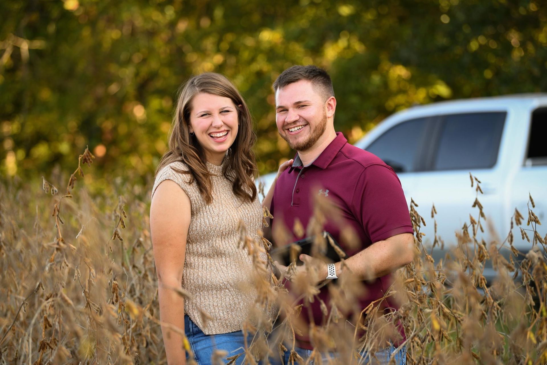 Woman with long brown hair smiles, arms crossed, wearing a plaid shirt, standing outdoors with green and yellow background.