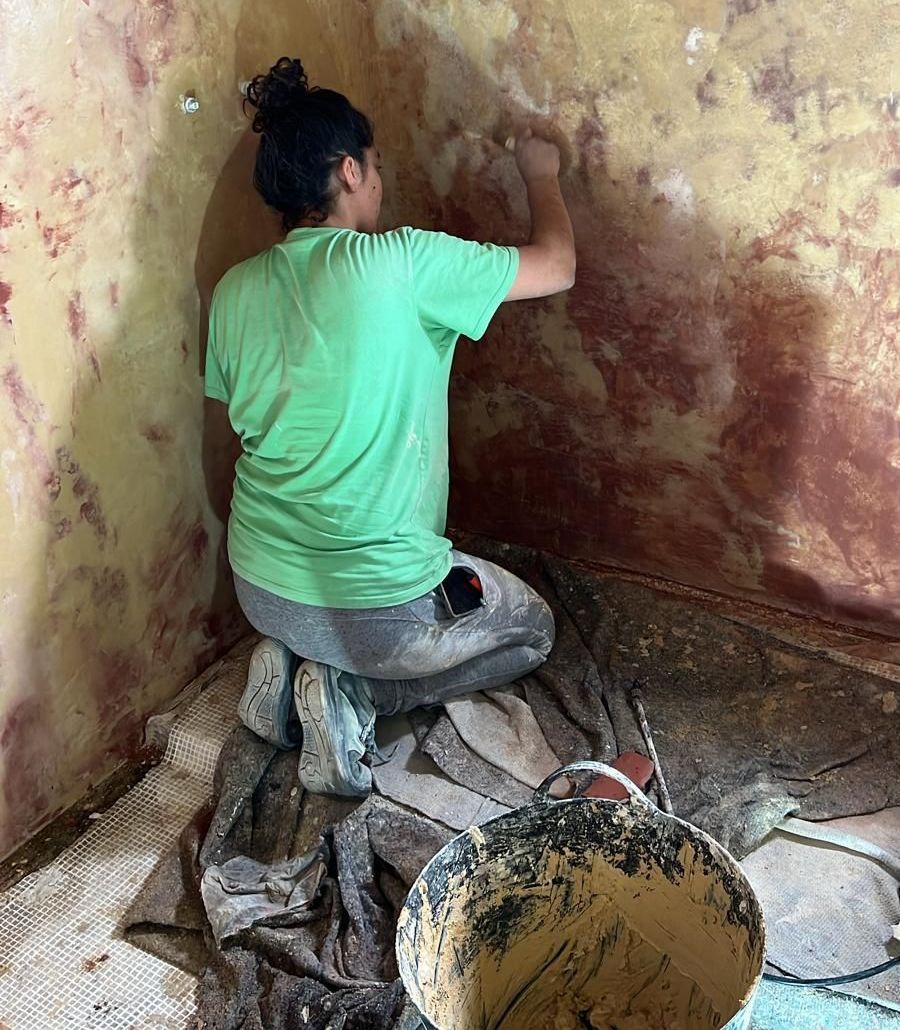 Una mujer con una camisa verde está arrodillada y pinta una pared.