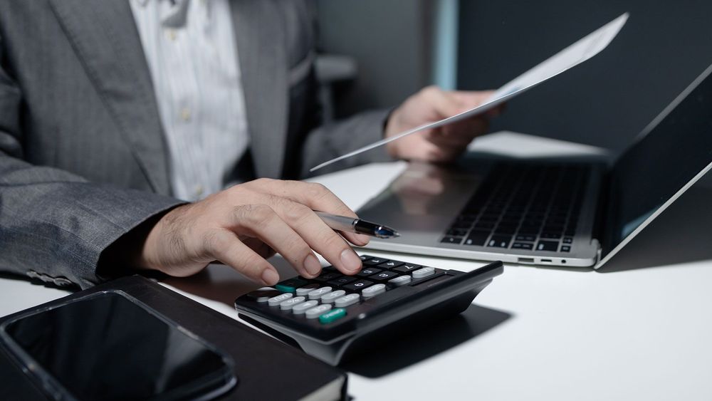 A man is sitting at a desk using a calculator and a laptop.