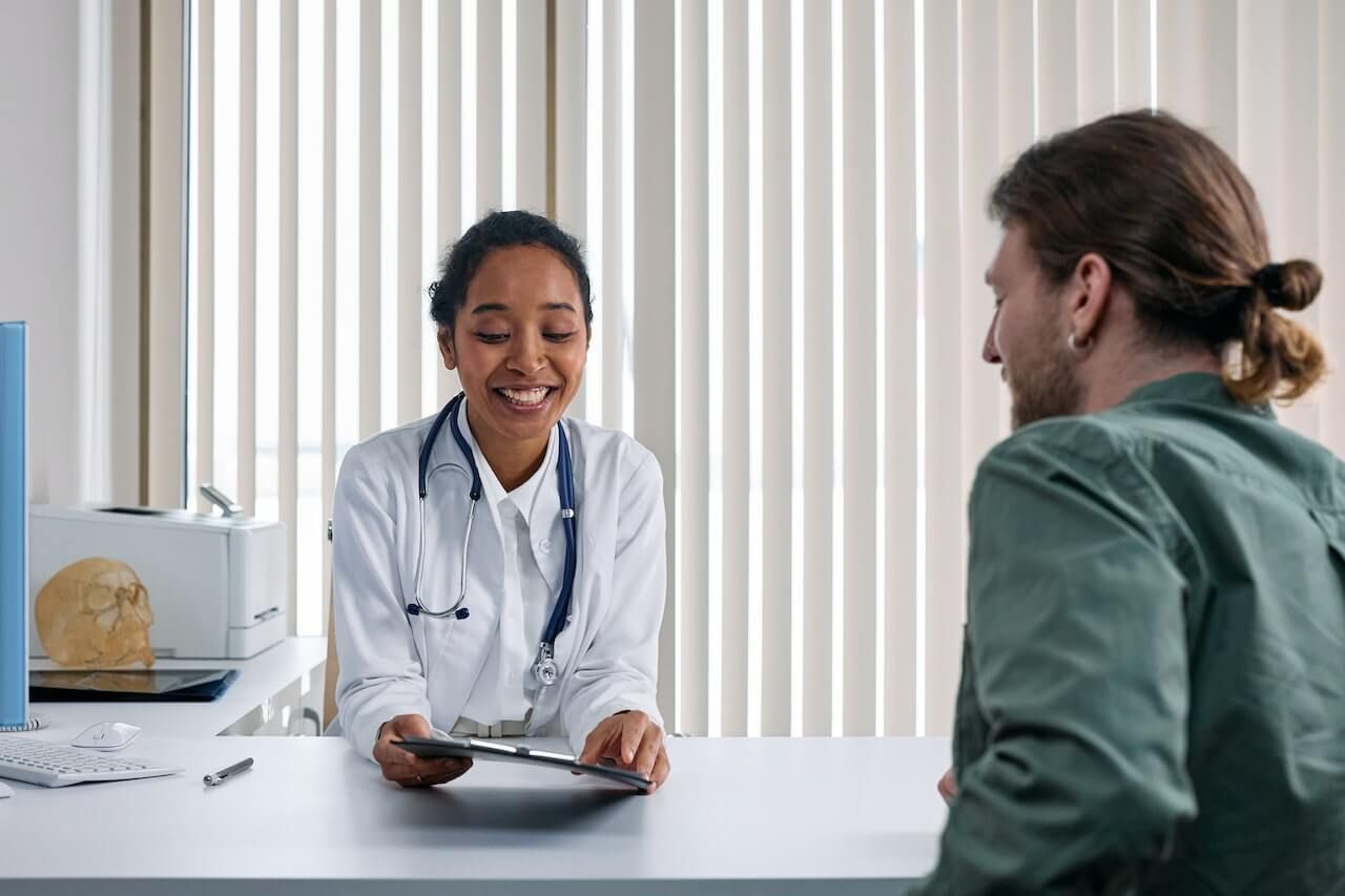 Doctor smiling, reviewing chart with patient in office. White coat, stethoscope, bright setting.
