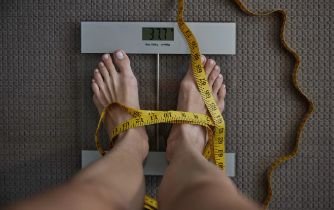 Feet on scale, wrapped with yellow measuring tape; digital display shows weight.