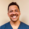 Smiling man with a mustache wearing blue scrubs in a room with a beige wall.