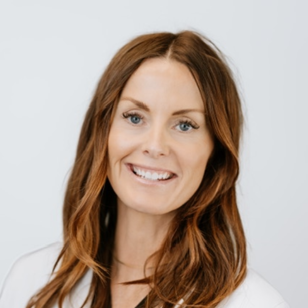 Woman with auburn hair smiles, wearing a white coat, against a white backdrop.