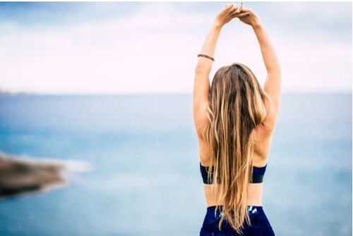 A woman with long hair stands on the beach, arms raised, celebrating wellness and vitality at a medical care clinic.