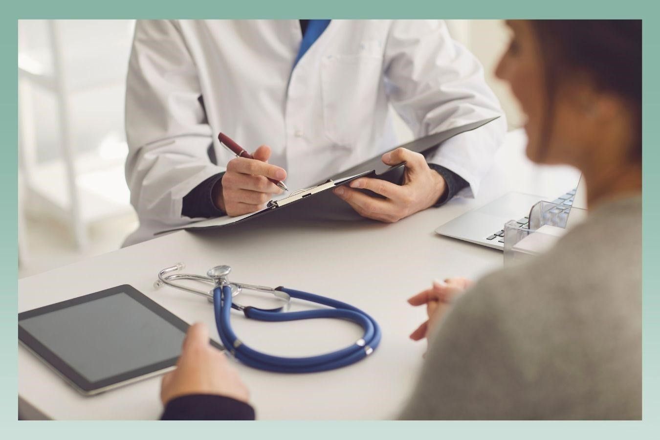 Doctor taking notes, speaking with a patient at a desk, stethoscope, tablet present.