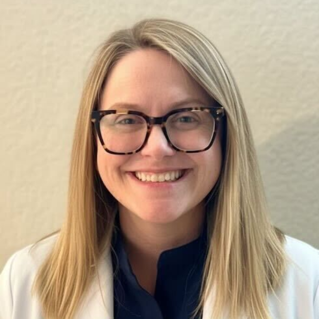 Woman with short blonde hair smiles in a white coat, outdoors in front of a building.