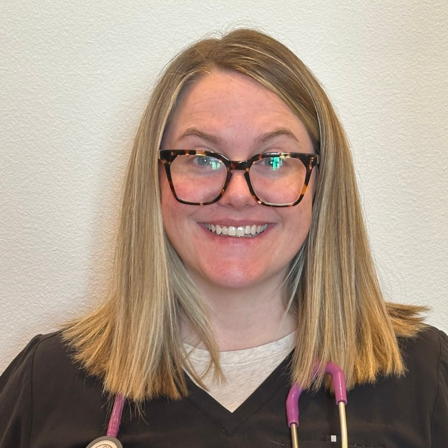 Woman with short blonde hair smiles in a white coat, outdoors in front of a building.