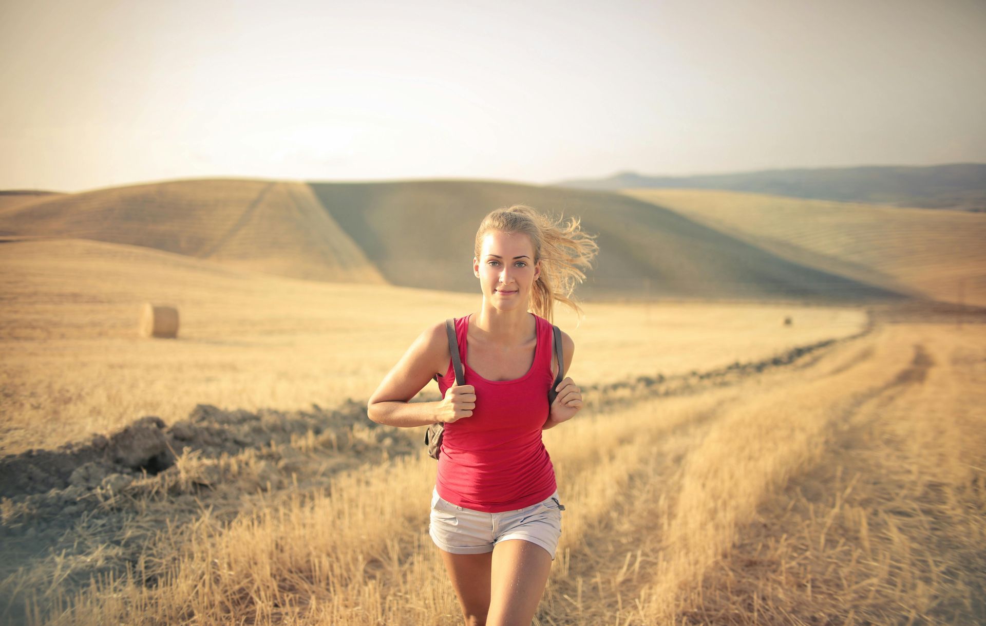 Blonde woman in red shirt and shorts runs on a dirt path through a golden field, wearing a backpack.