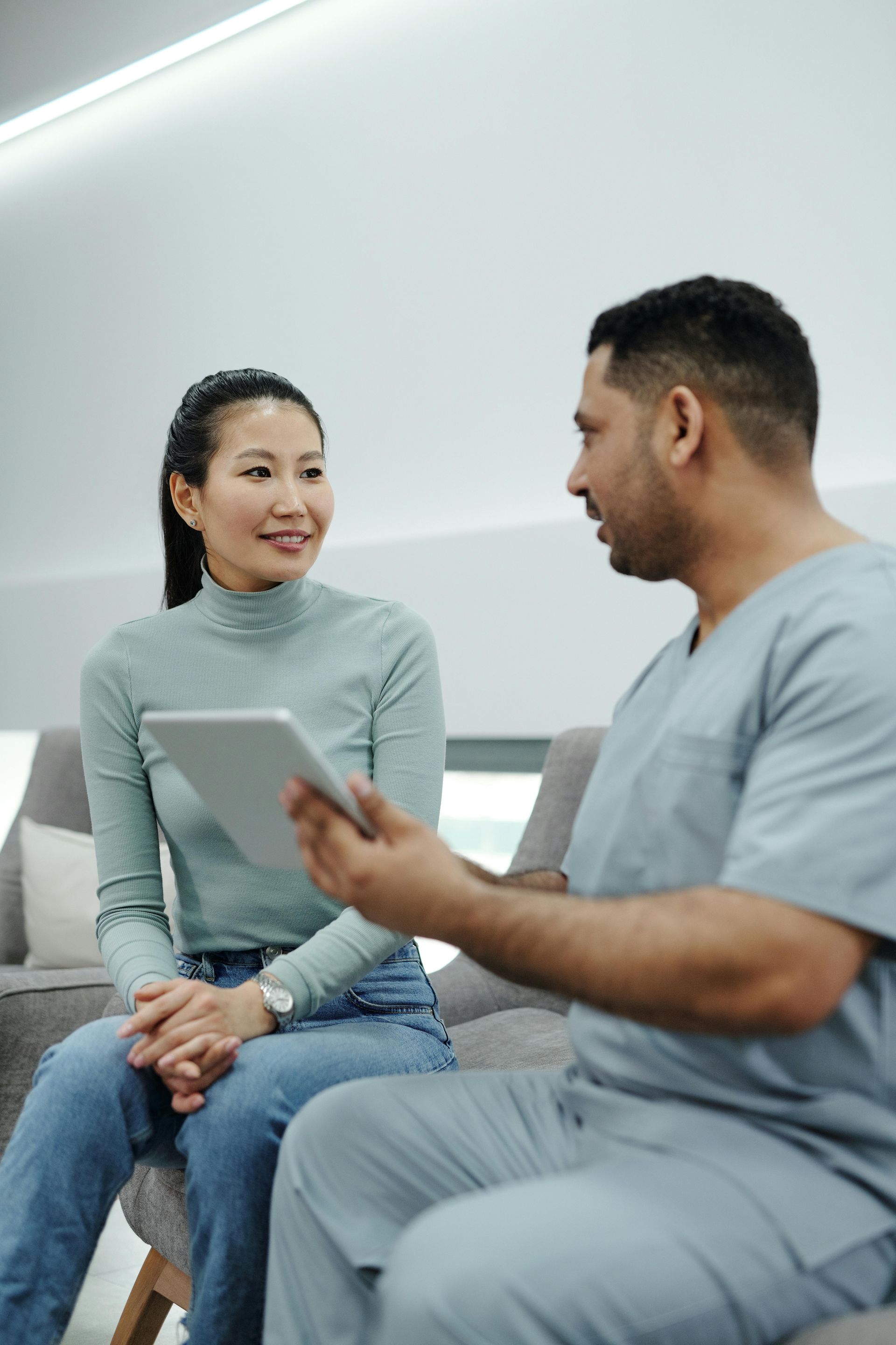 Woman seated, looks at medical professional holding tablet. They appear to be in a medical office setting.