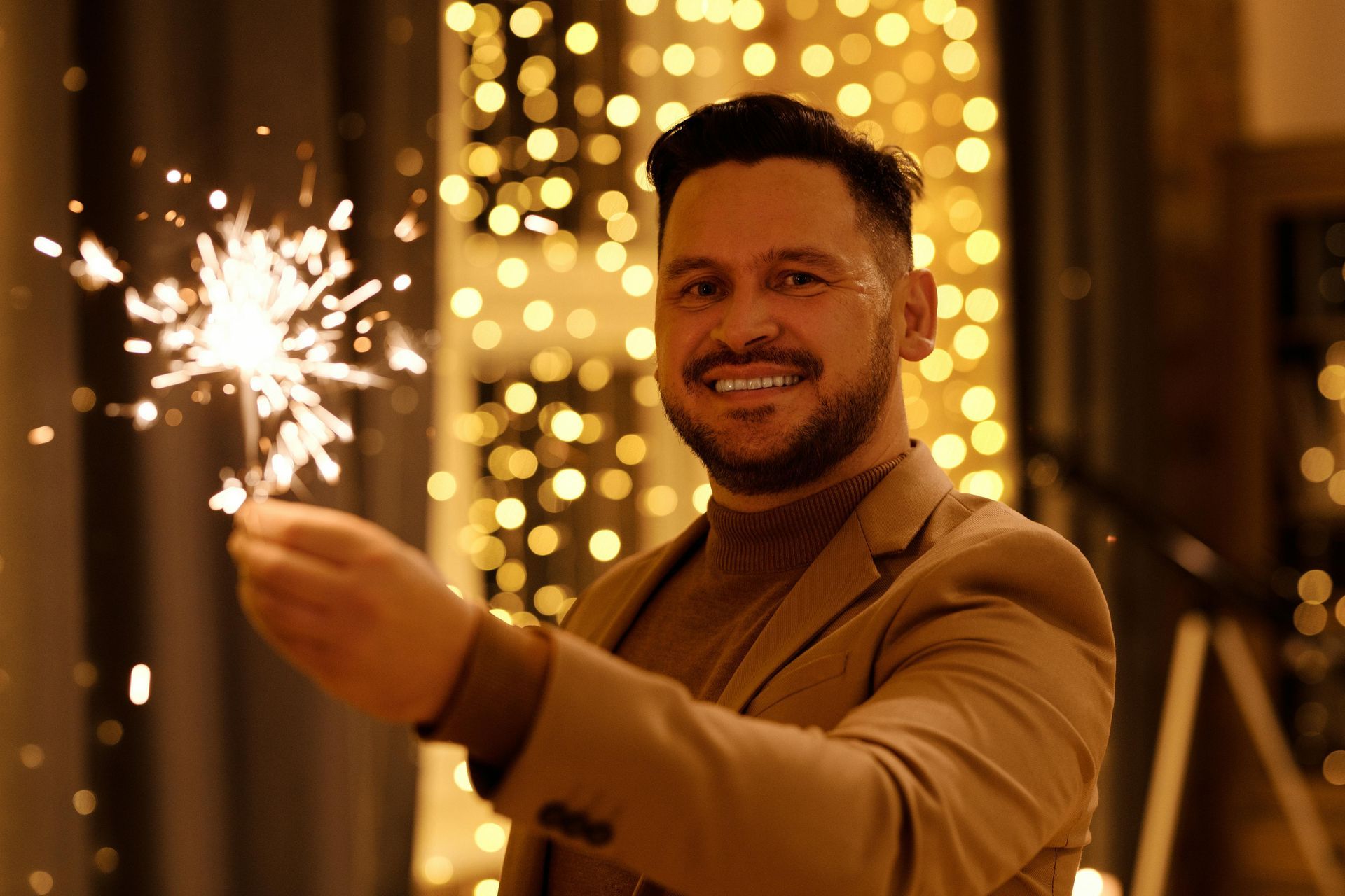 Man smiling, holding sparkler in front of a bokeh background with warm lighting.