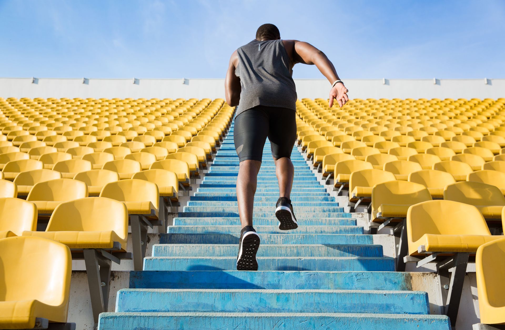 Man runs up stadium steps. Bright blue steps, yellow seats, clear sky.