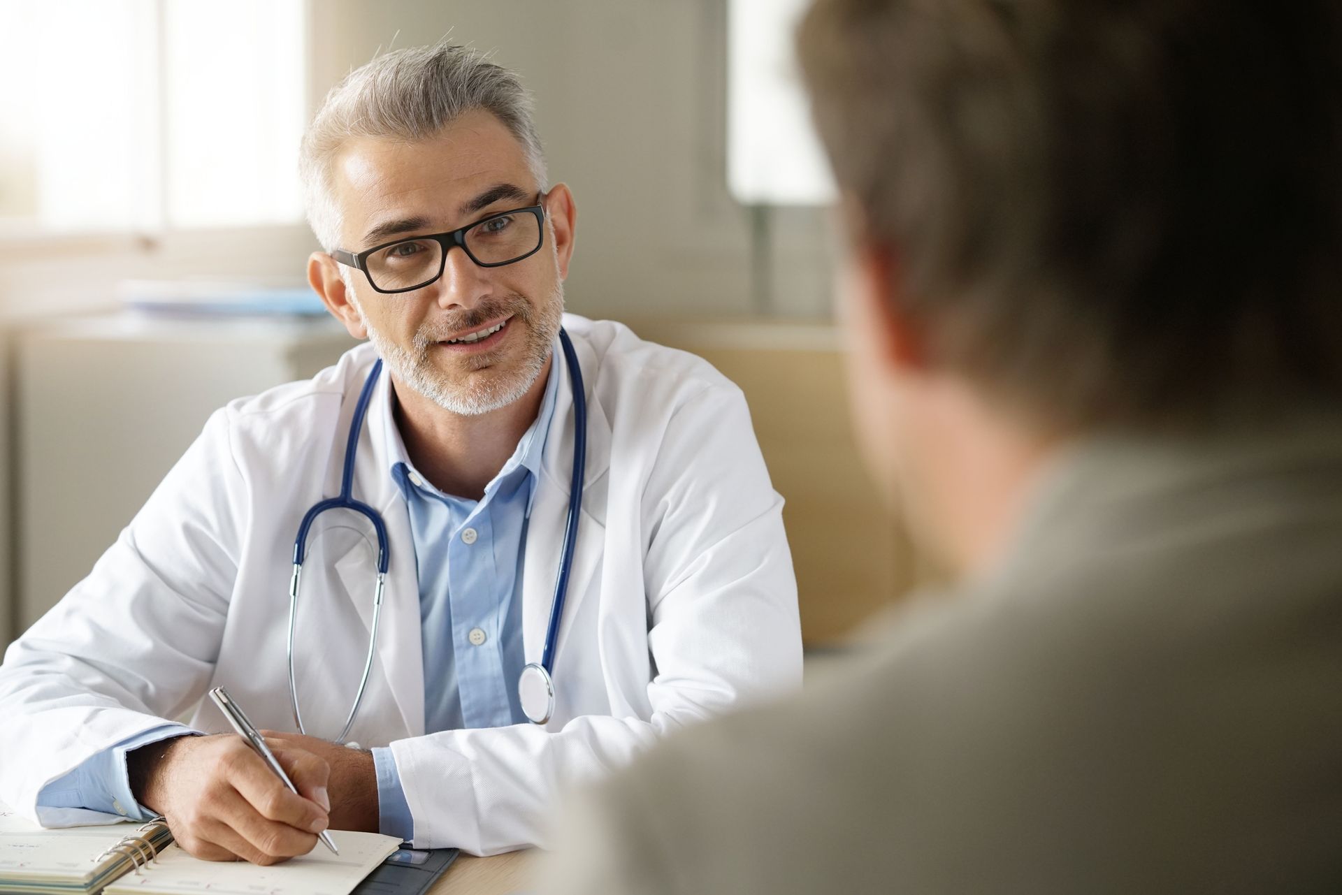 Doctor in white coat with stethoscope, smiling, talking to patient in office.