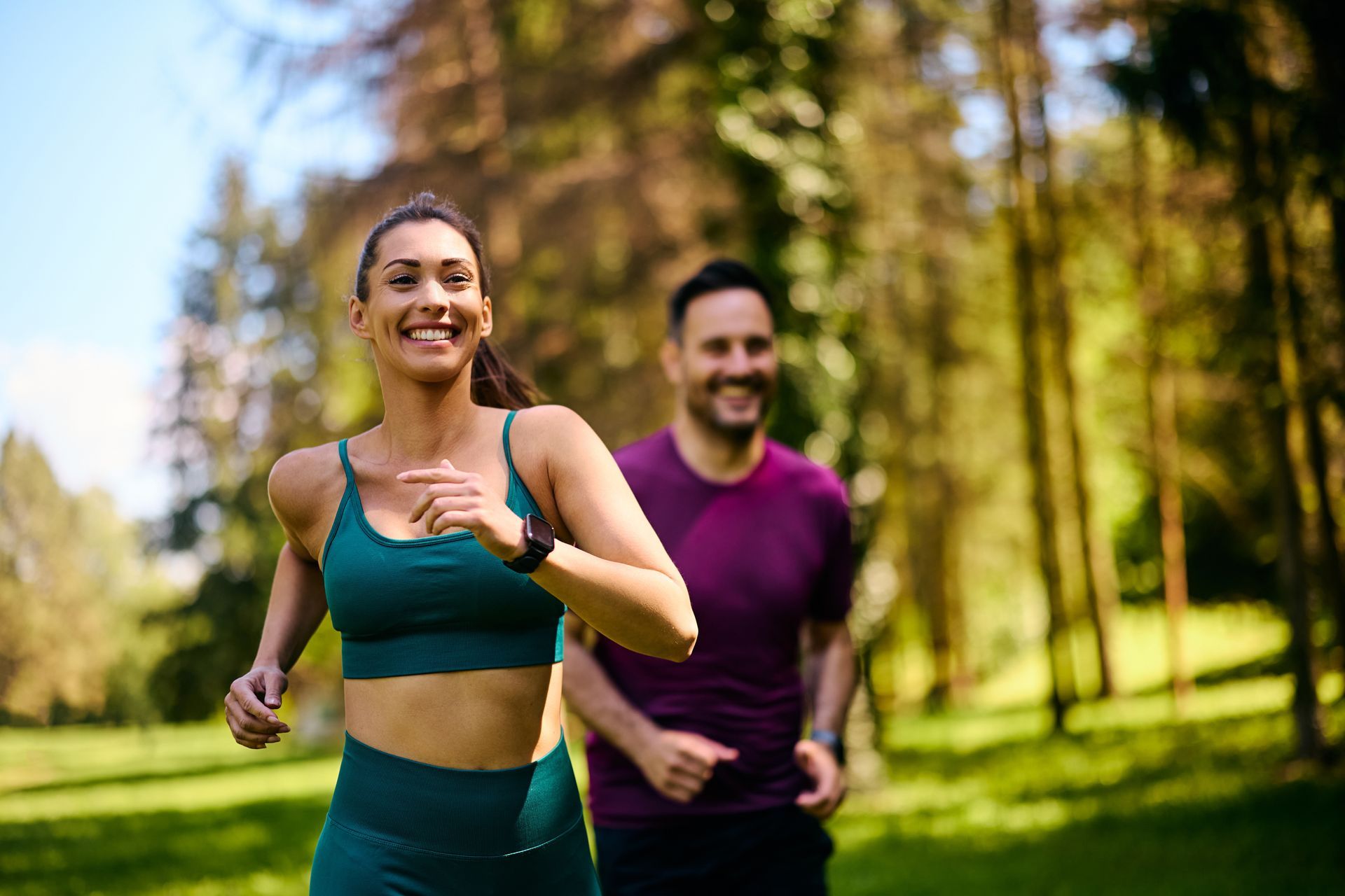 Woman and man running in a park, smiling. Woman in teal sports bra and leggings. Man in purple shirt. Sunny day.