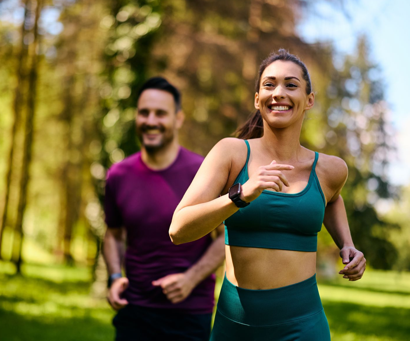 Woman and man smiling while jogging in a park; woman in teal sports bra, man in purple shirt.