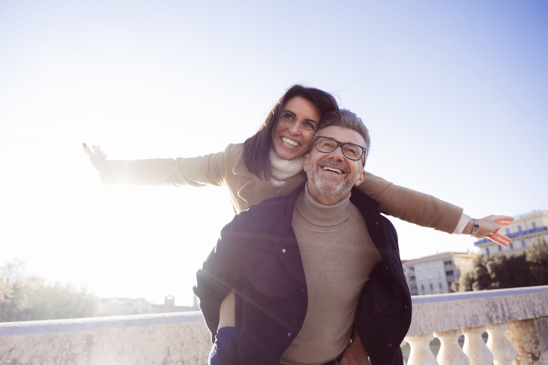 Smiling man giving woman a piggyback ride, arms outstretched, outdoors in sunlight.