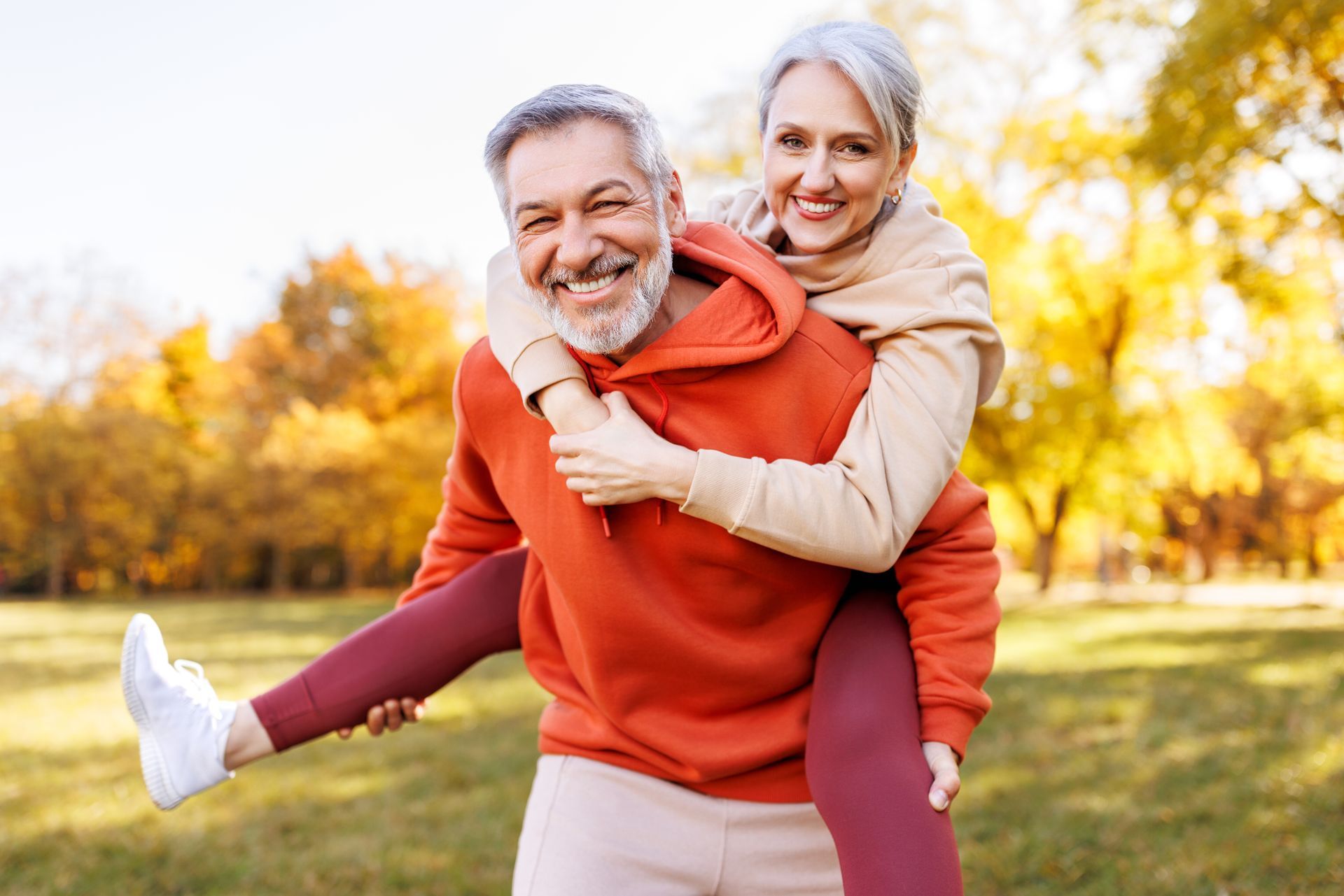 Smiling older couple, man giving woman a piggyback ride in a park with fall foliage.