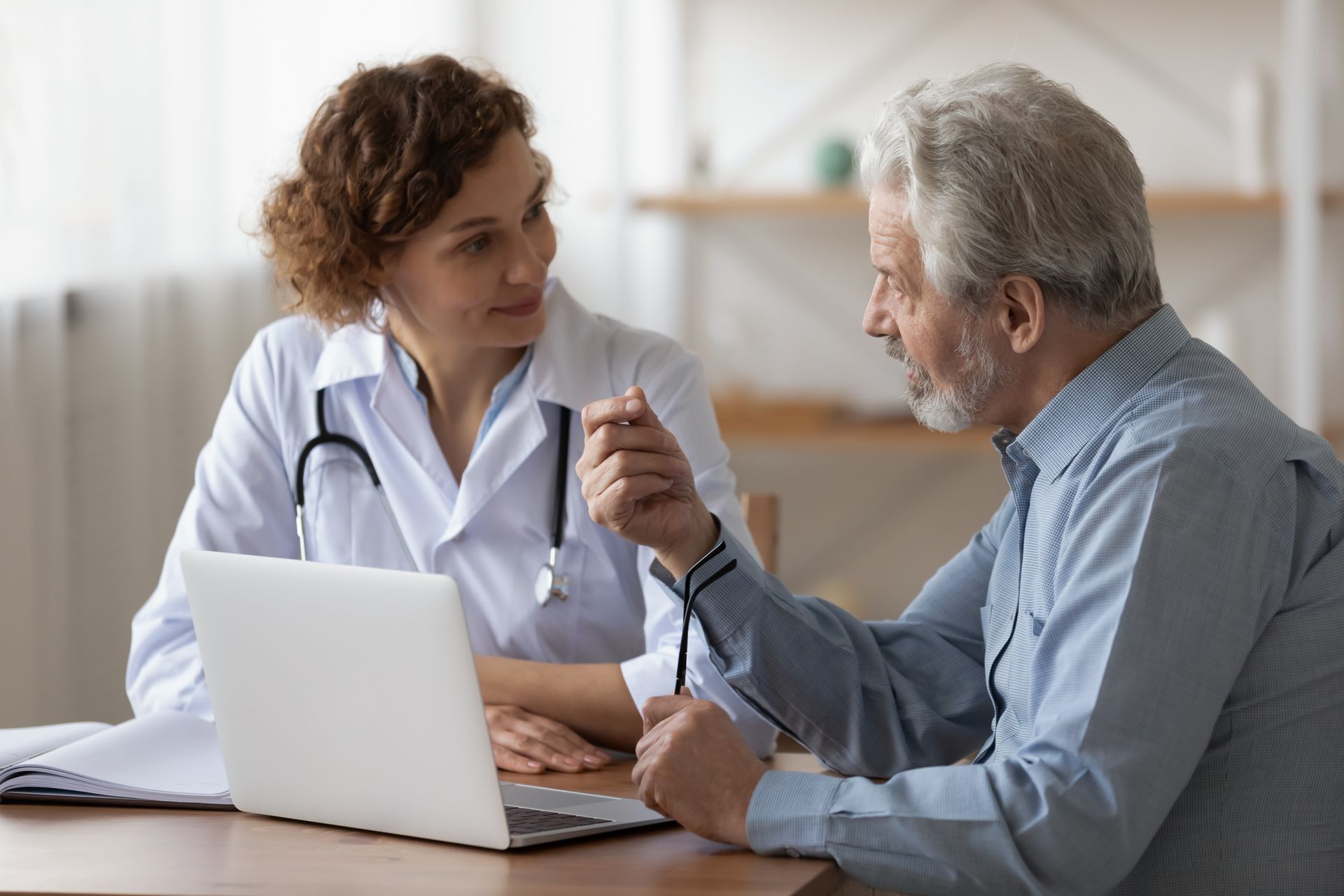 Doctor in white coat consults with elderly man at a table, a laptop between them.