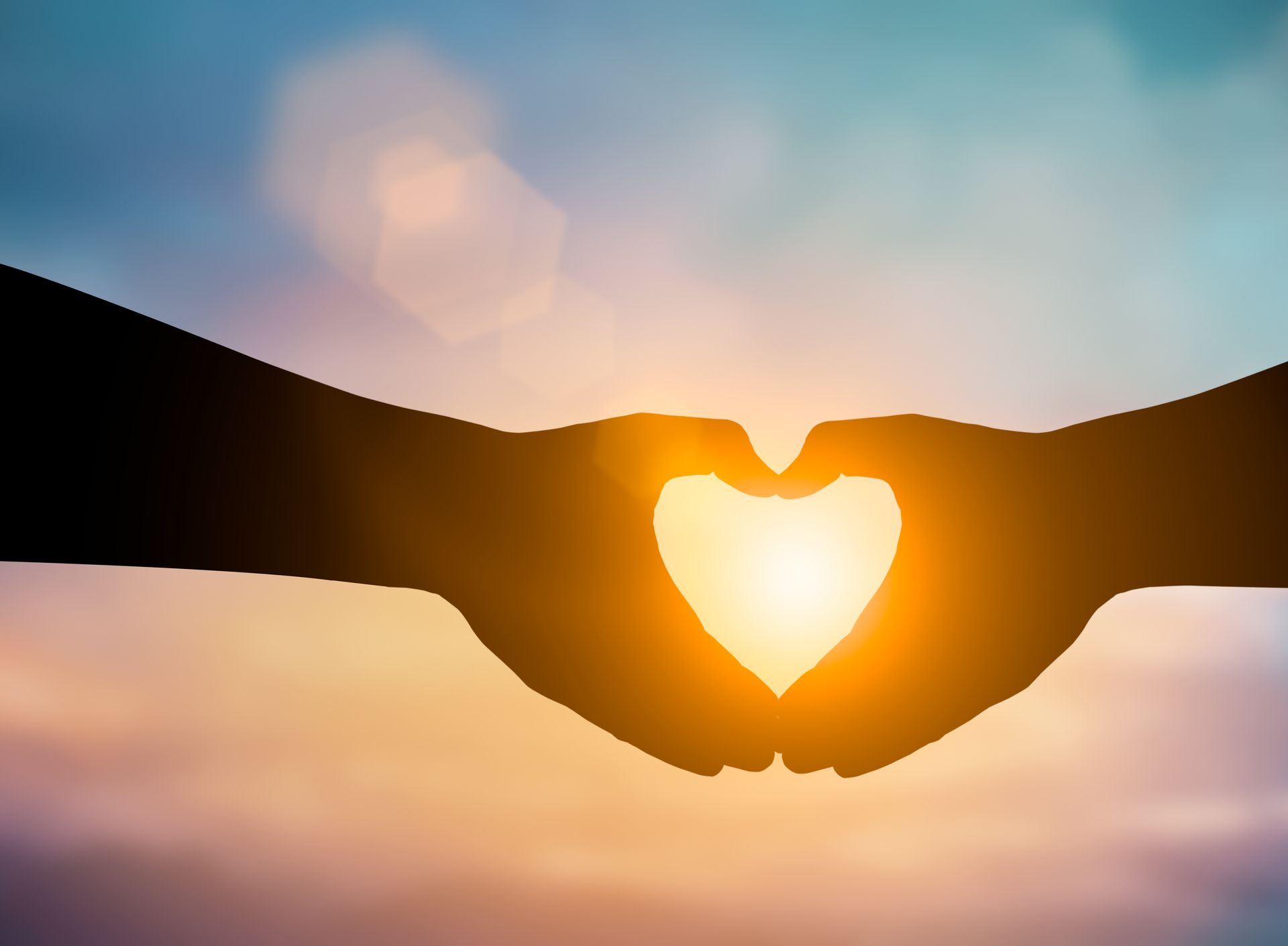 Silhouette of hands forming a heart shape, with bright sunlight shining through against a colorful sky.