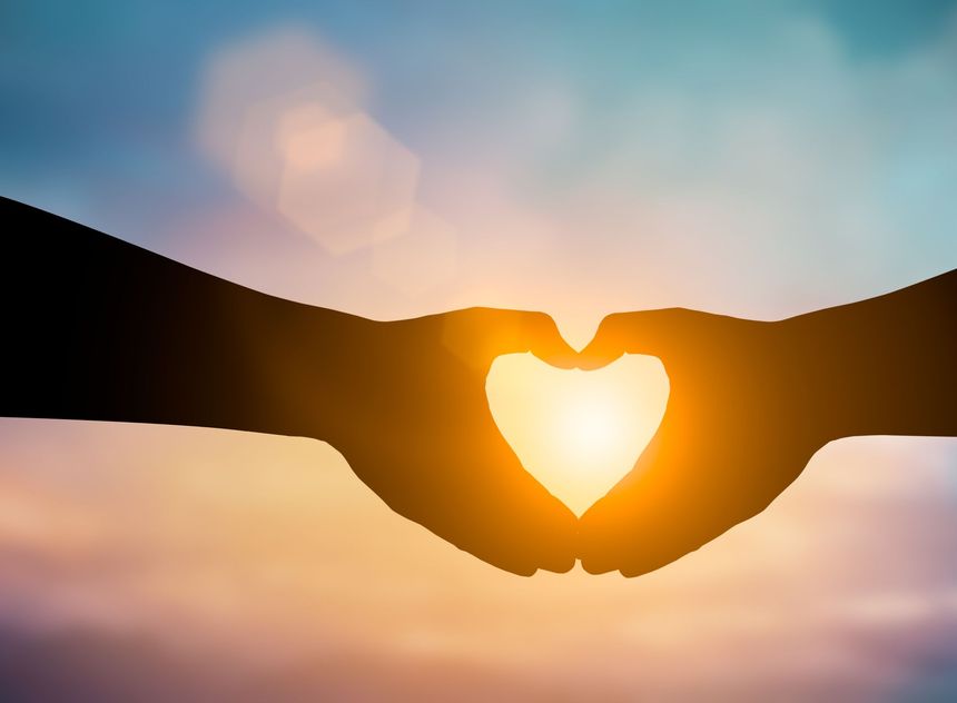 Silhouette of hands forming a heart shape, with bright sunlight shining through against a colorful sky.