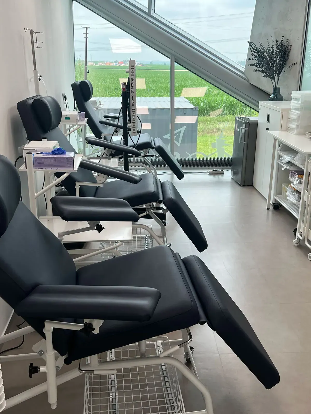 Rows of reclining chairs in a medical facility, next to a window overlooking green fields.