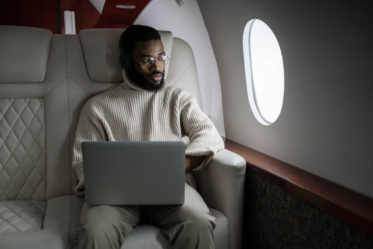Black man in a cream sweater, using a laptop on a private jet, looking out the window.
