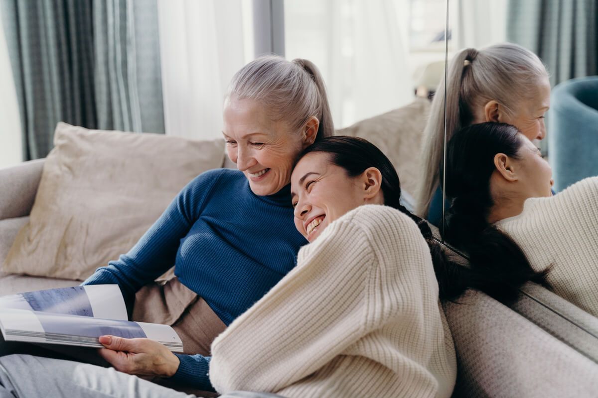 Two women of different ages, smiling and embracing on a sofa while looking at a photo album.
