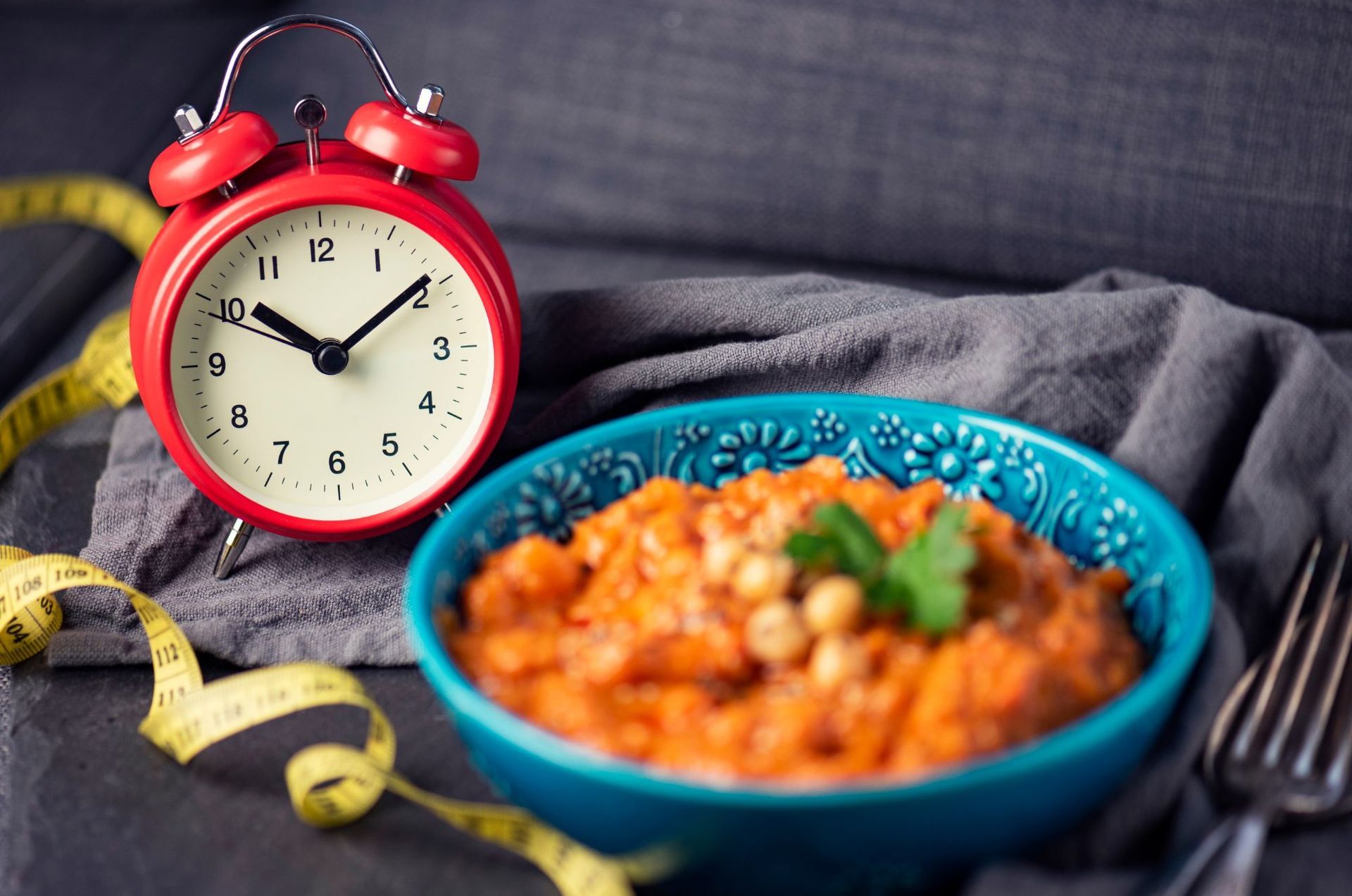 Red alarm clock, teal bowl of food, measuring tape, and fork on gray cloth.