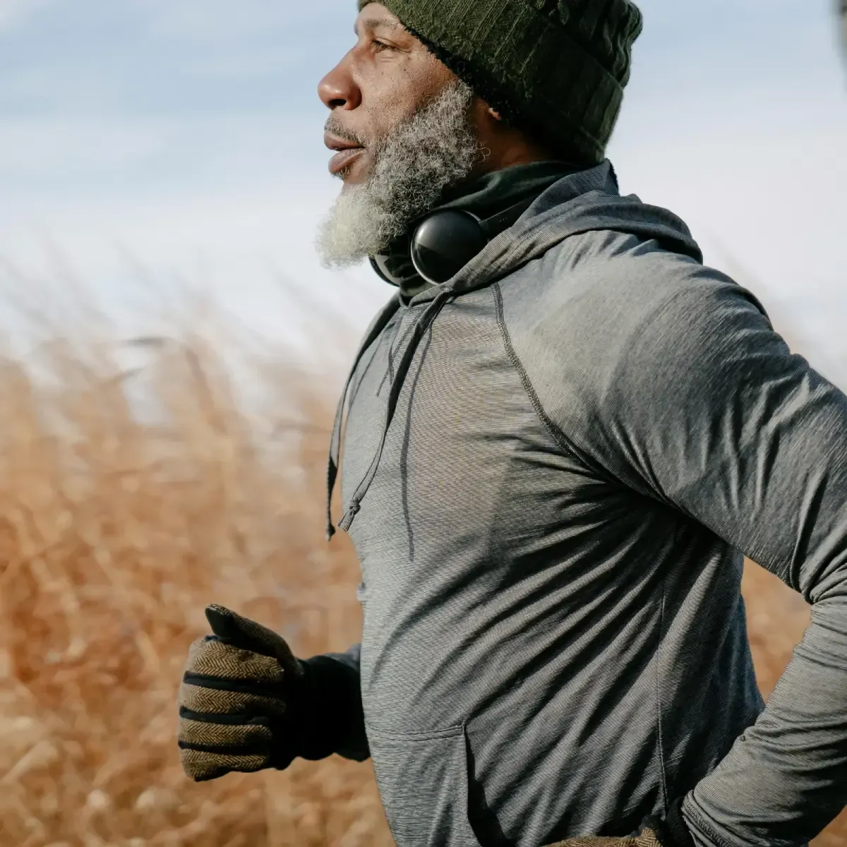 Black man in grey hoodie, green hat, and gloves running outdoors.