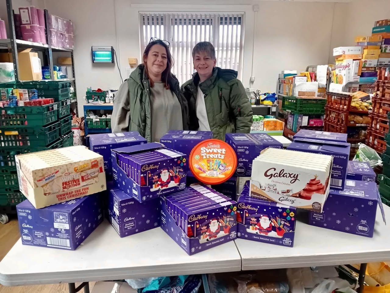Two people stand behind a table piled with chocolate boxes and a tin. They are in a room with shelves of food.