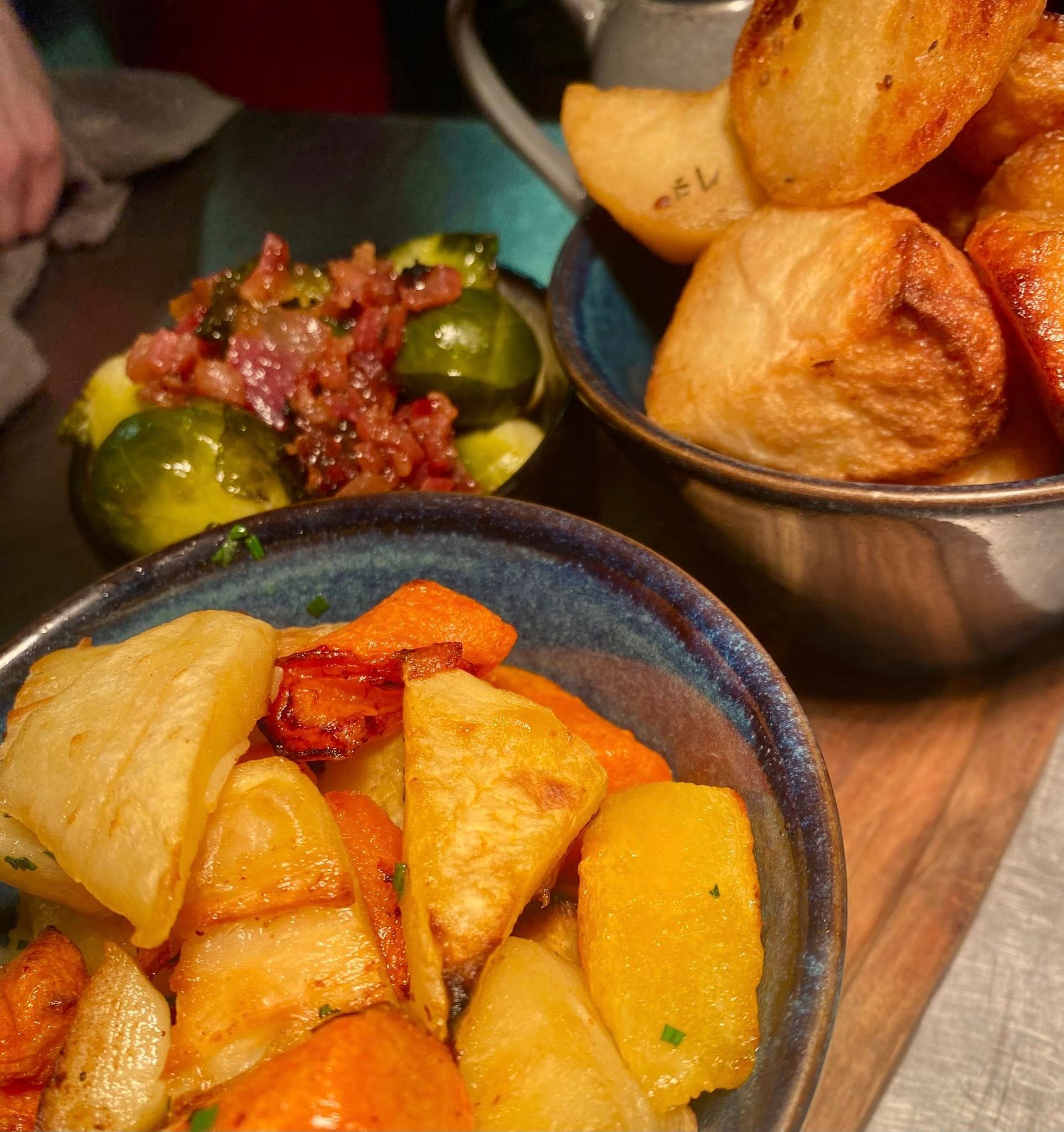 Bowls of roasted vegetables: potatoes, carrots, and Brussels sprouts with bacon, on a wooden board.