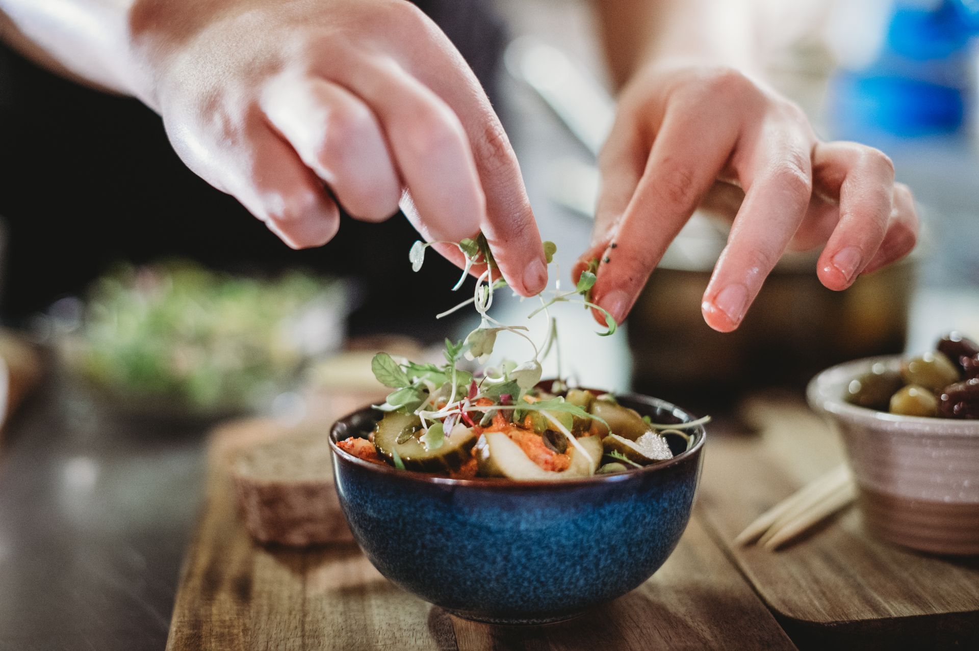 Hands garnishing a blue bowl of salad with sprouts on a wooden cutting board.