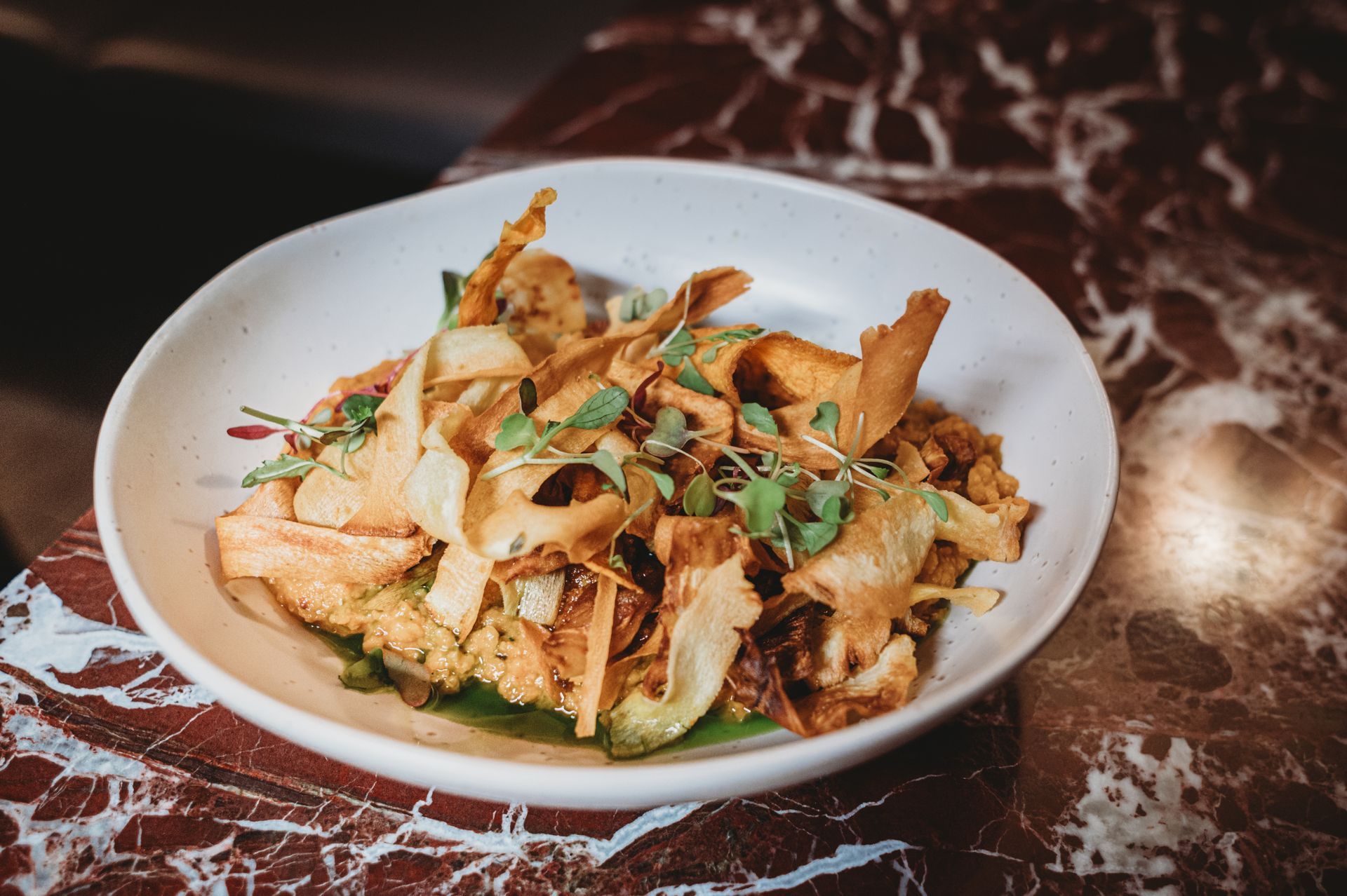 Close-up of a white bowl with colorful food, including crispy chips and greens, on a marble table.