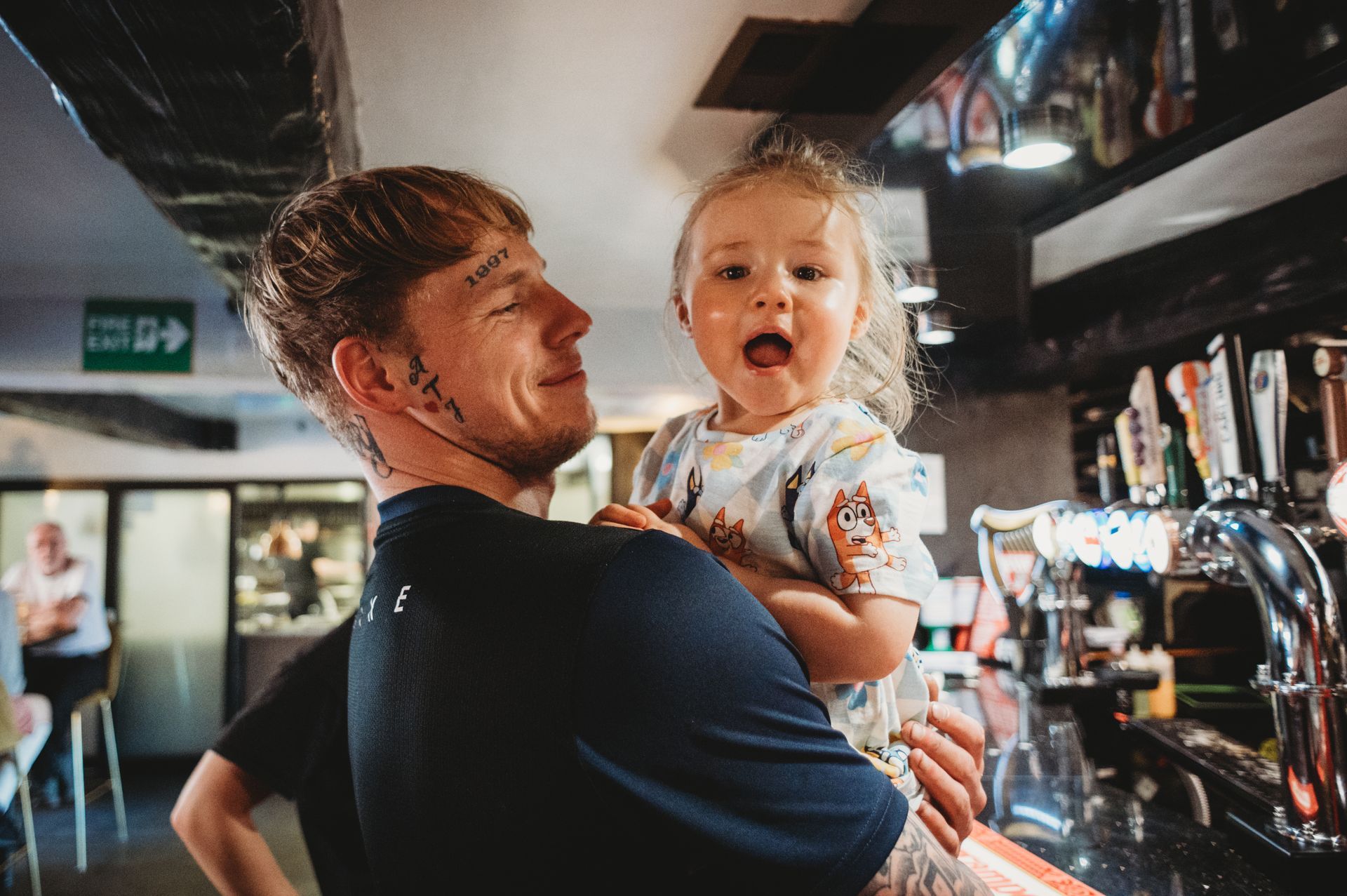 Man holding a child at a bar. Both looking towards the camera; the child with mouth open.
