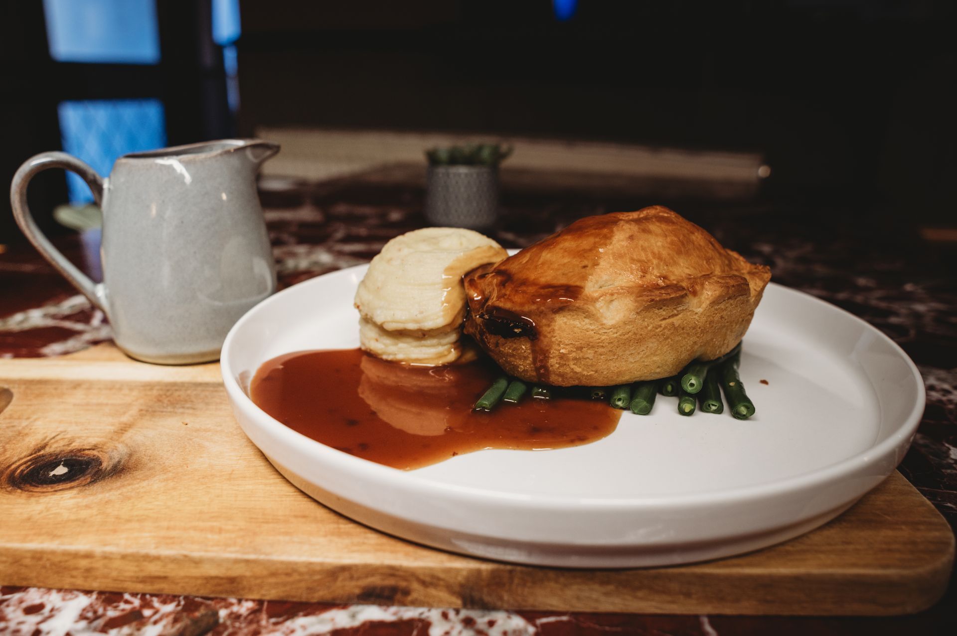 Meat pie with gravy, mashed potatoes, and green beans on a white plate, wooden board, gravy boat.