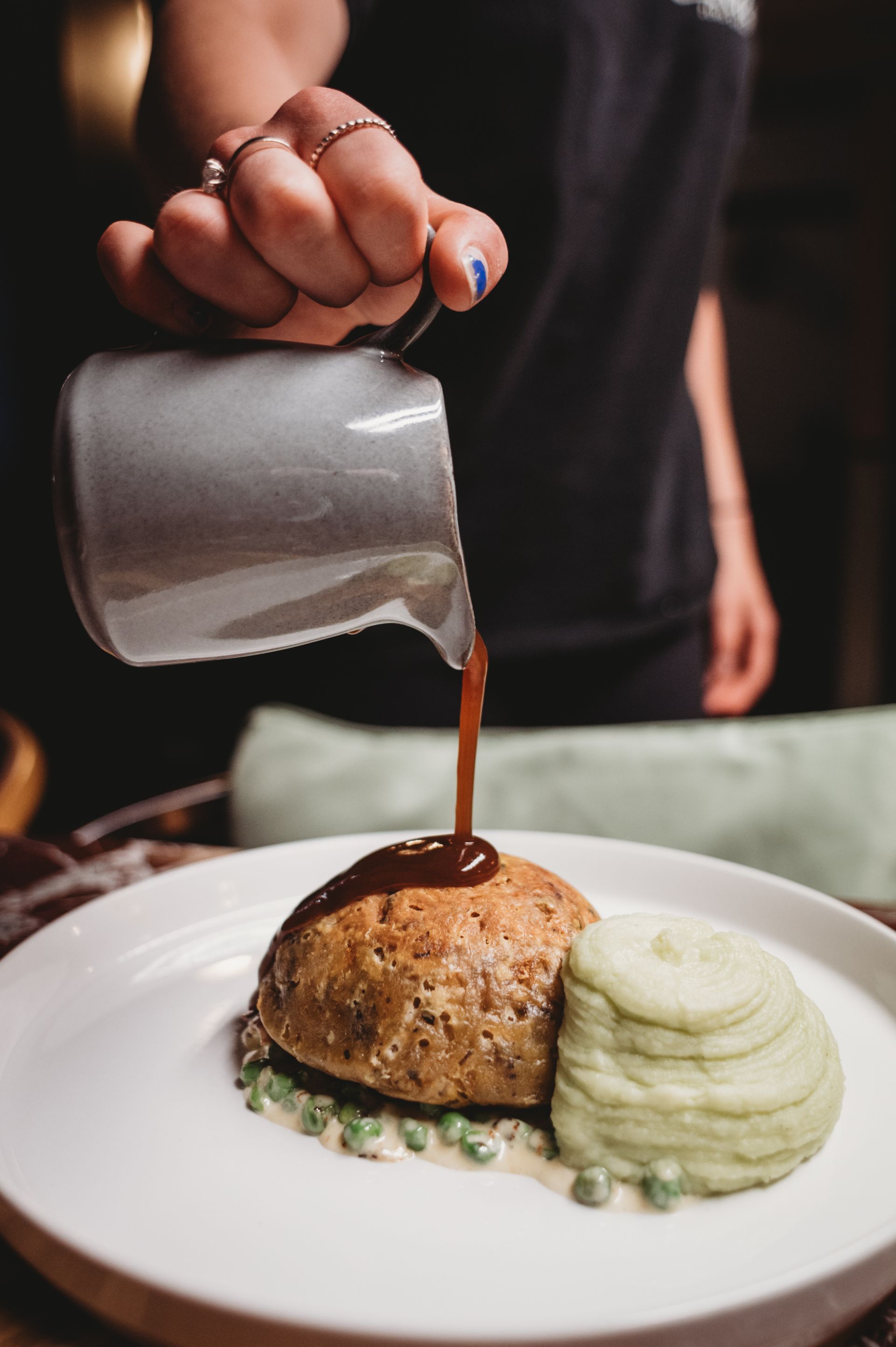 Gravy being poured over a meat pie, served with mashed potatoes and peas on a white plate.