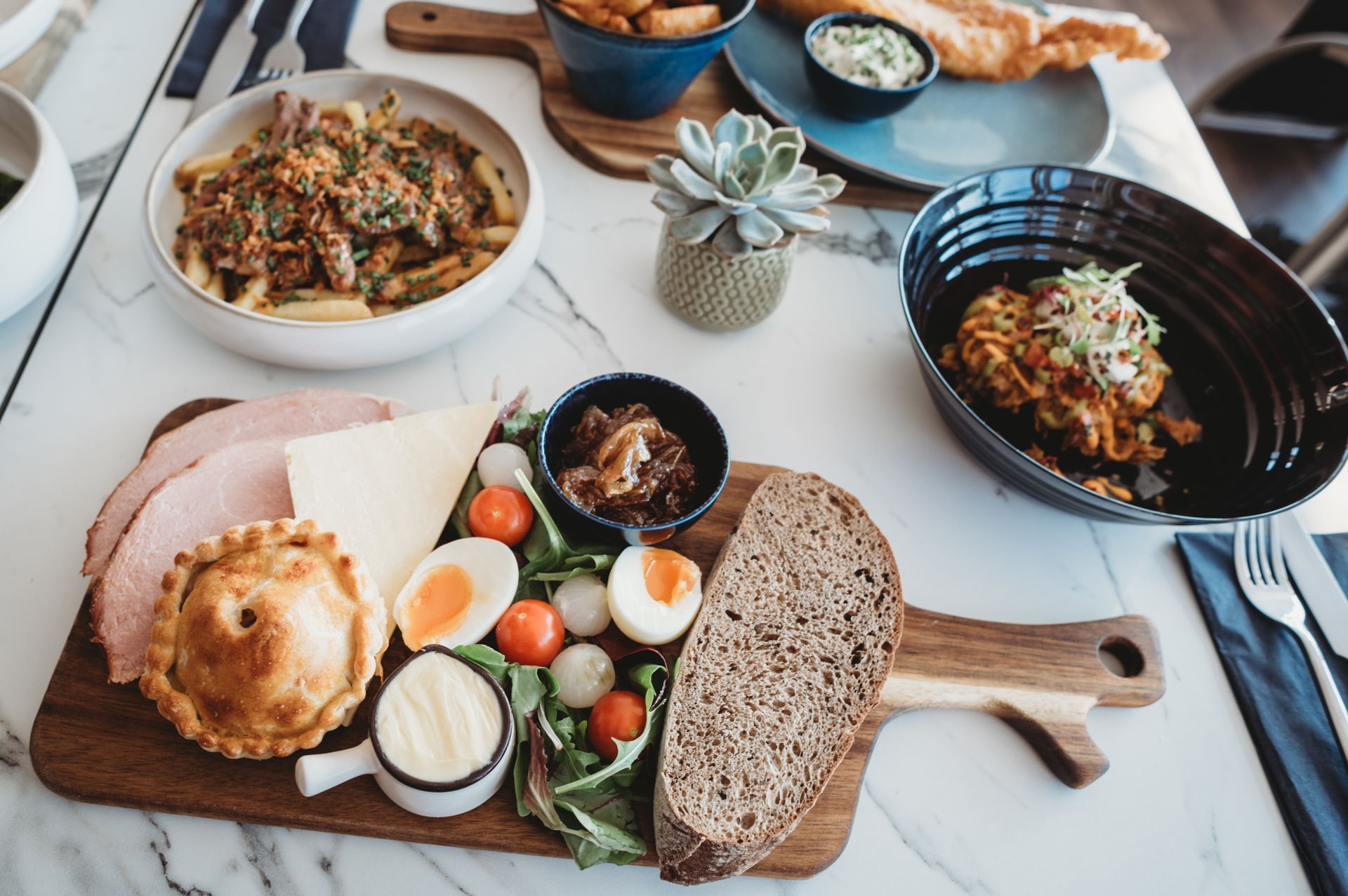 Wooden board with assorted food: pie, bread, cheese, eggs, salad, and other dishes on a marble-topped table.