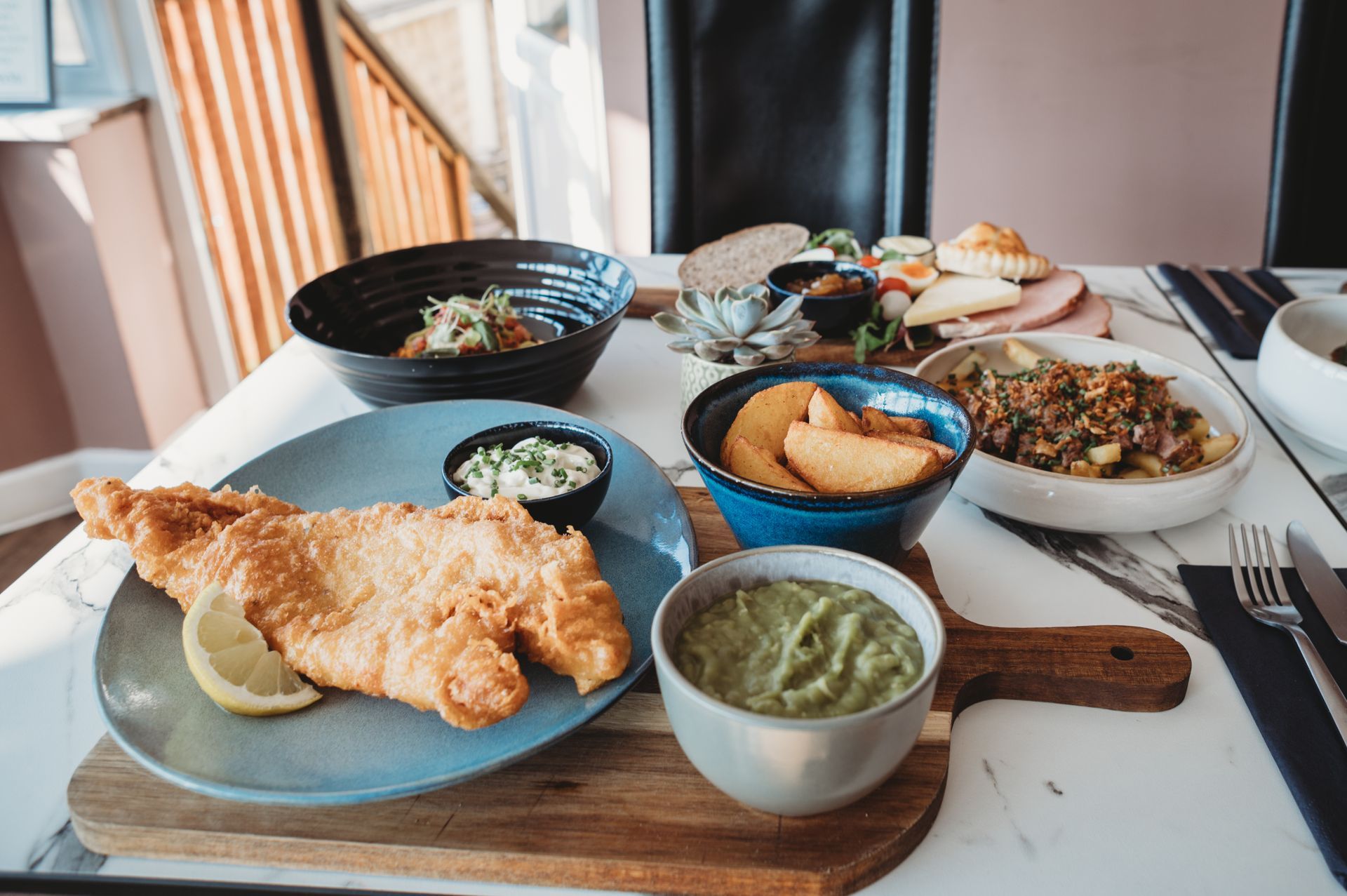 Fish and chips with sides, avocado dip, and assorted dishes on a marble-top table.