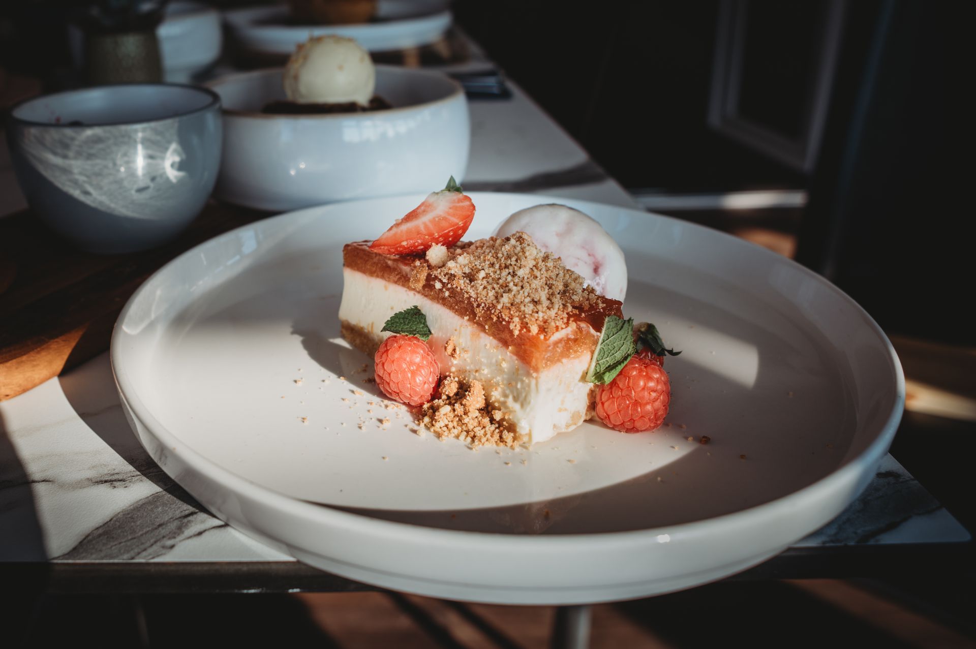 Cheesecake with strawberries, ice cream, and crumbs on a white plate in sunlight.
