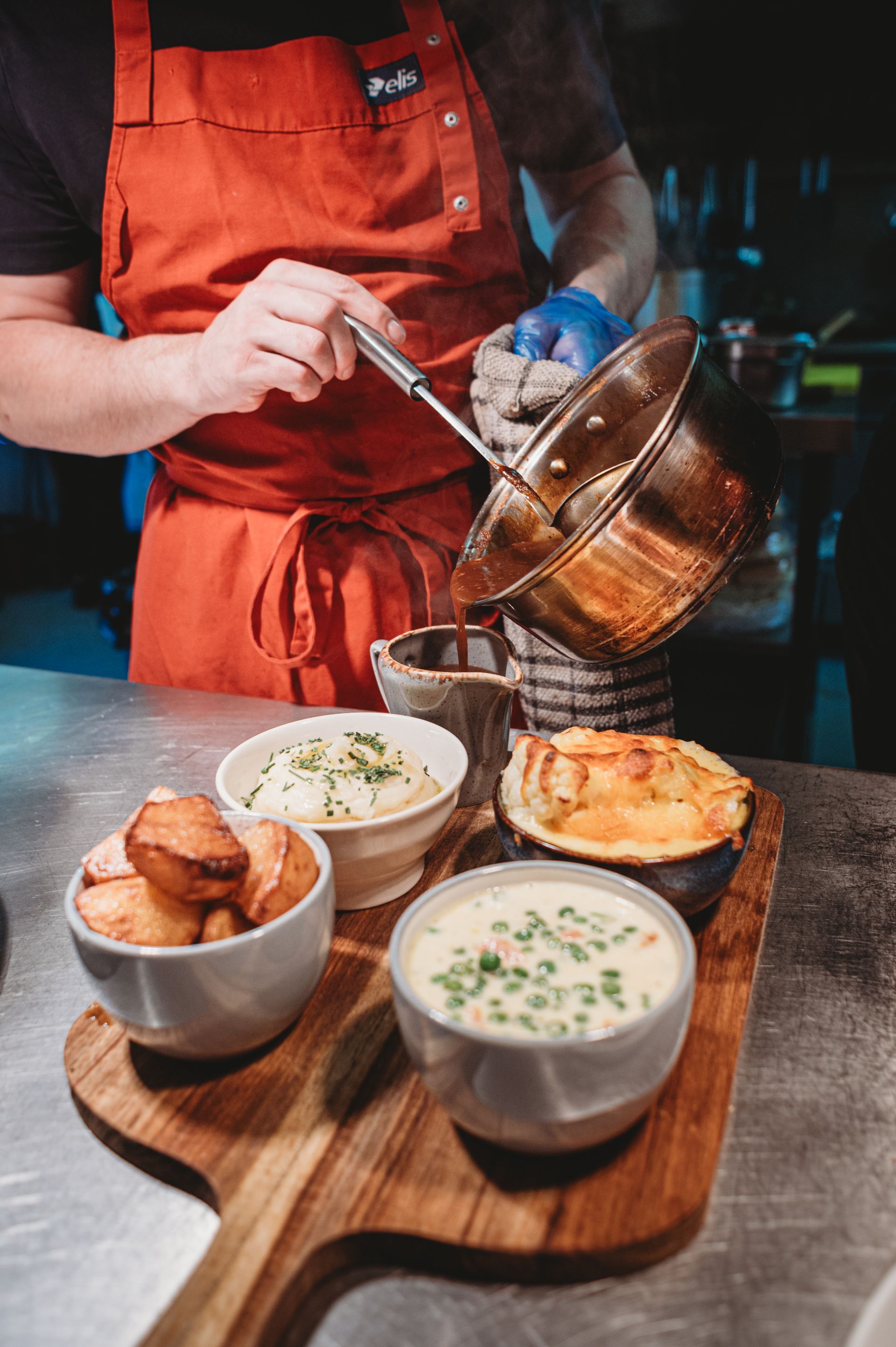 Chef pouring sauce over a platter of side dishes: potatoes, mashed potatoes, and a baked dish.