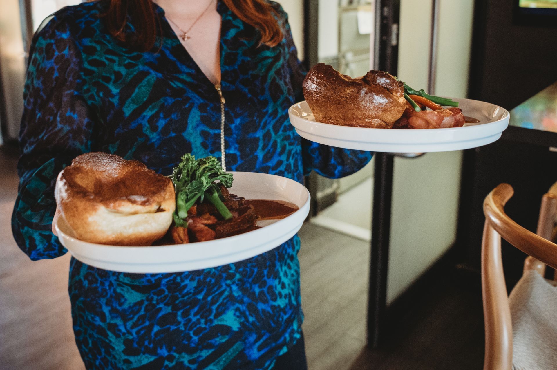 Woman in blue dress carries two plates of roast beef with Yorkshire pudding and vegetables.