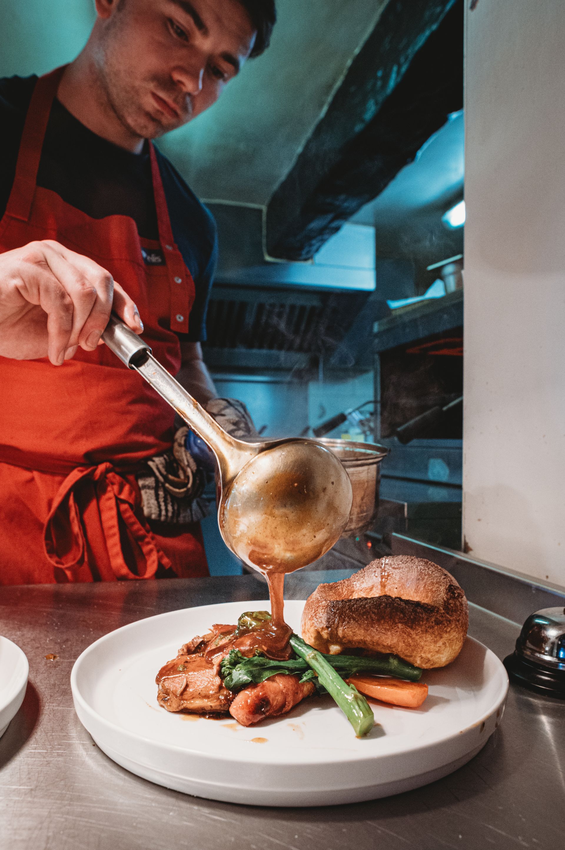 Chef pours gravy from a ladle onto a plate of roast beef, vegetables, and a Yorkshire pudding.