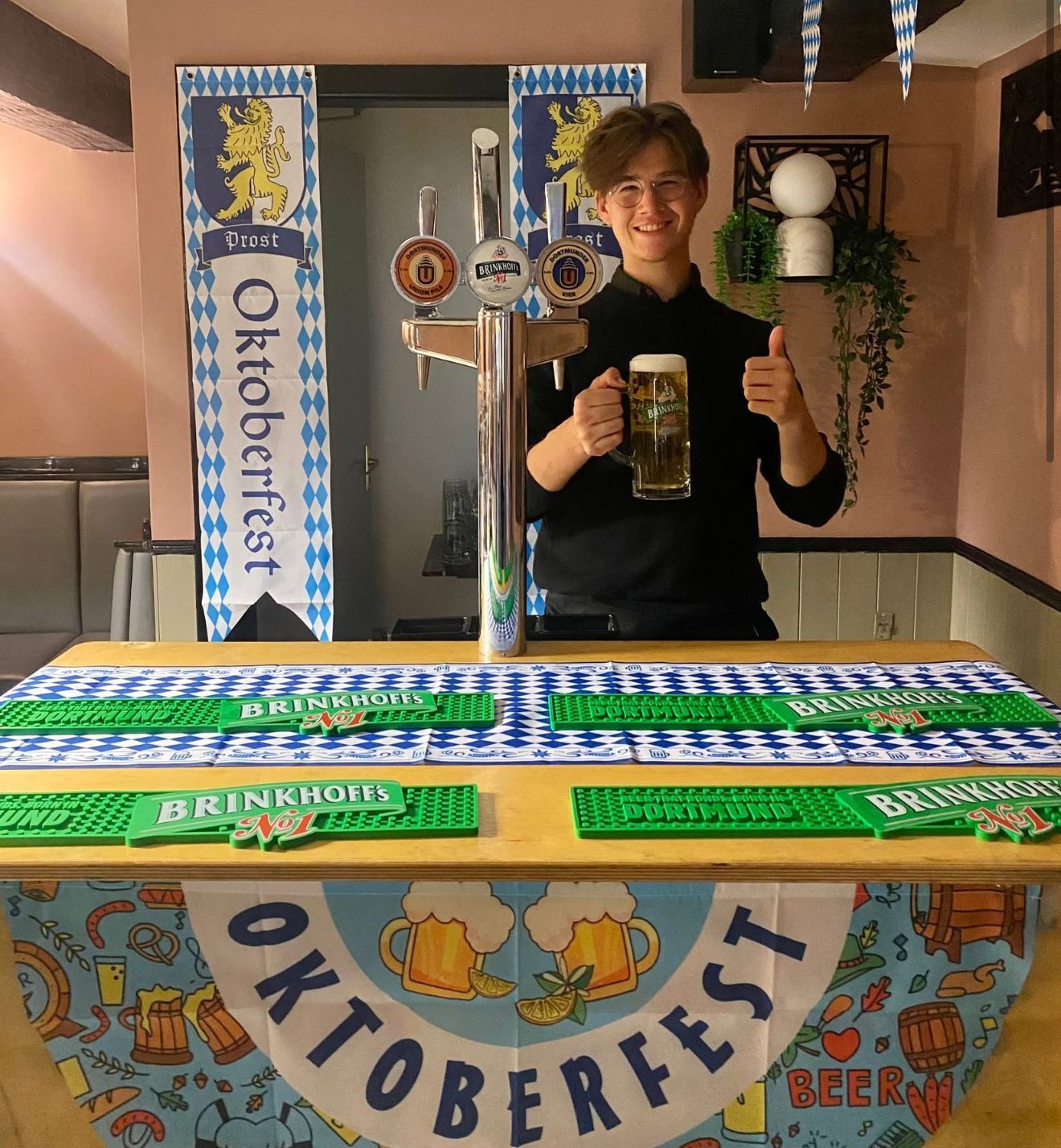 Bartender holding beer, giving thumbs-up Oktoberfest-themed bar with beer taps and banners.Boot & Shoe, Waterfoot, Rossendale