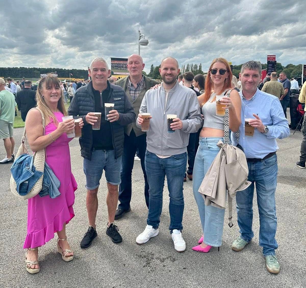 A group of six people smiling, holding drinks, outdoors on a cloudy day.