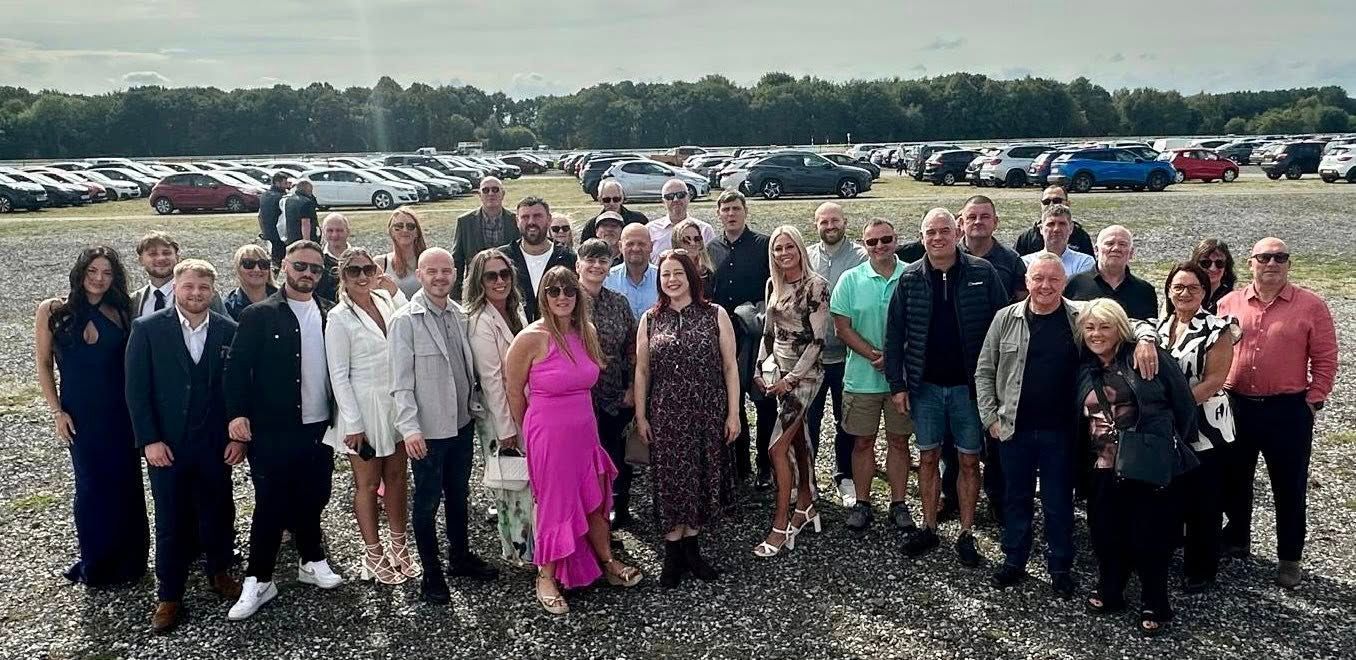 Group of people posing outdoors in front of cars and trees. Cloudy sky.