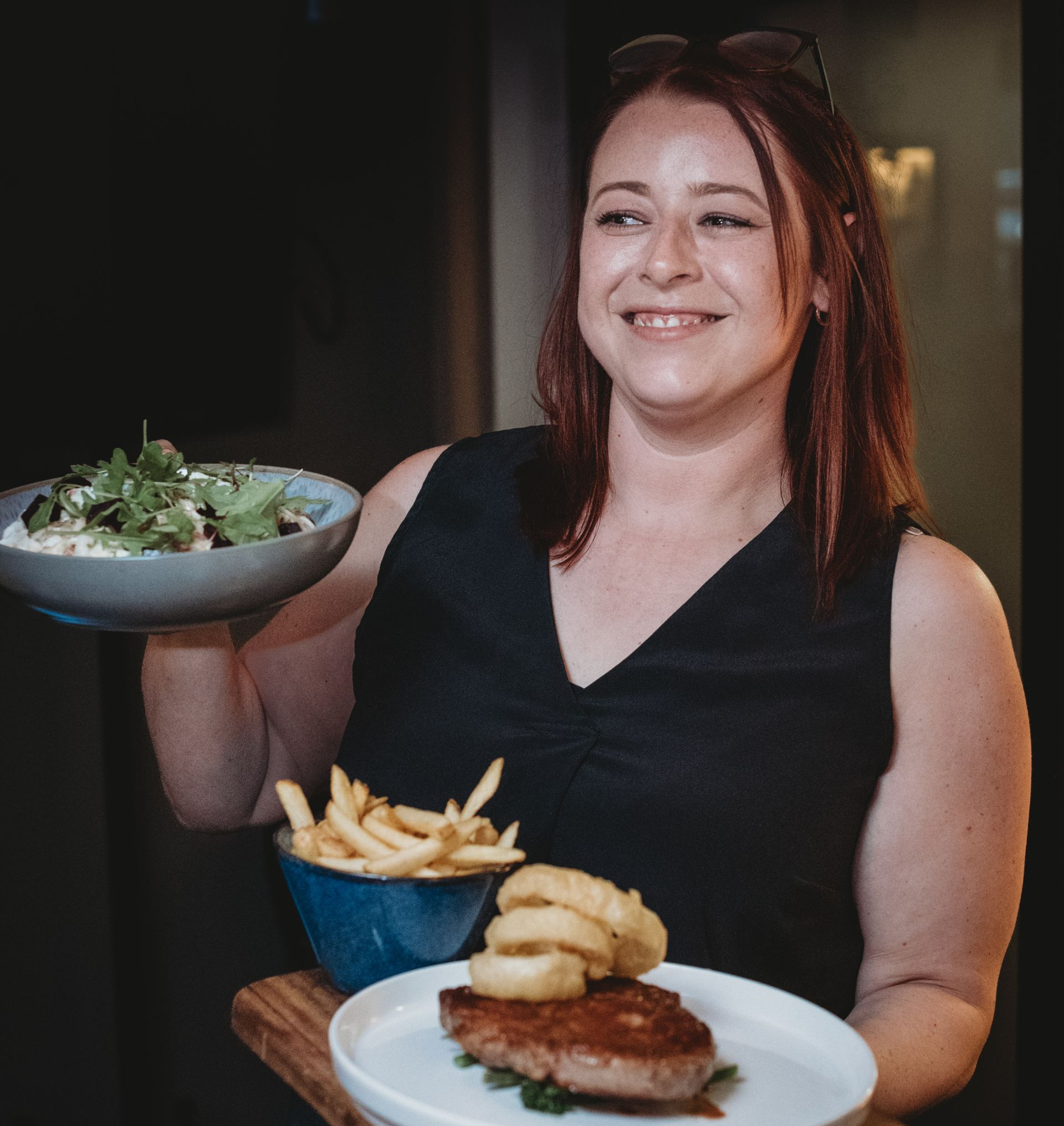 Woman smiles, holding plates with food: pasta, fries, steak, and onion rings. at the Boot & Shoe Waterfoot, Rossendale