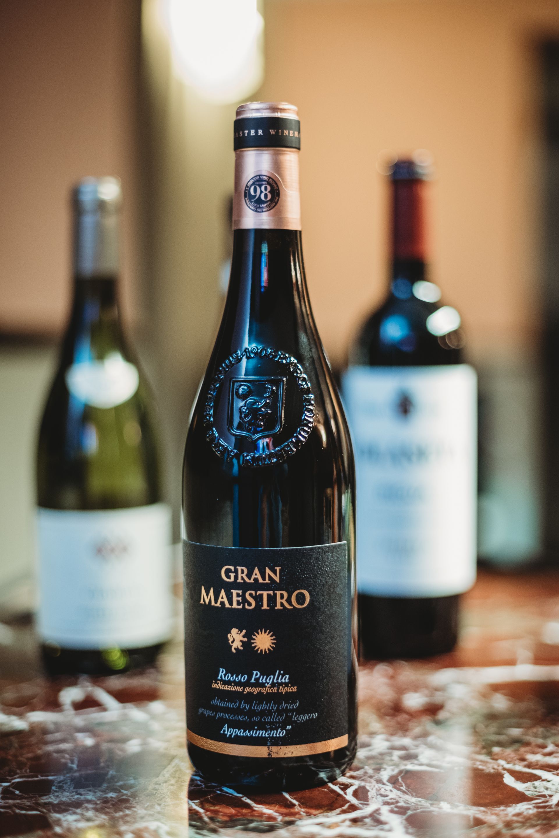 Three wine bottles on a table. Center bottle is Gran Maestro, black label, burgundy wine. Other two bottles blurred.