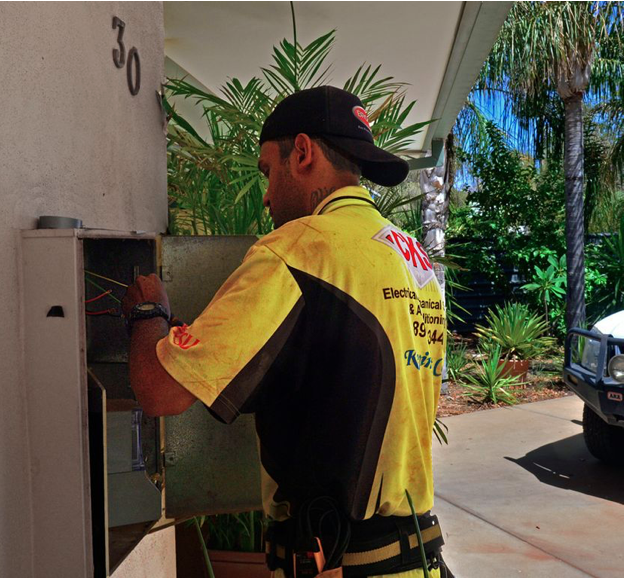 Electrician in yellow shirt working on a grey electrical box outside a house with palm trees. — CKS Electrical and Air Conditioning in Ciccone, NT