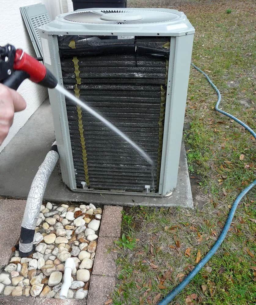 Person Cleaning an Air Conditioning Unit With a Water Hose Outdoors — CKS Electrical and Air Conditioning in Yulara, NT